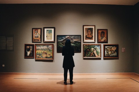Das Foto zeigt eine Szene im Museum. Auf einer grauen Wand sind verschiedene Gemälde angebracht. Davor steht mittig im Bild eine Person in dunkler Kleidung. Die Person betrachtet die Bilder.