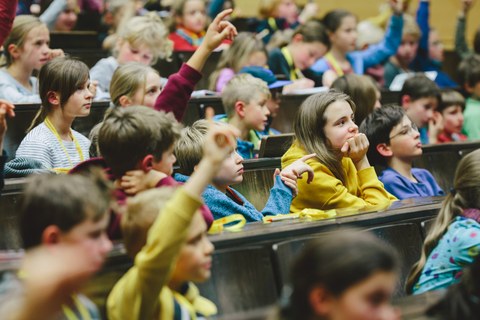 In einem Hörsaal sitzen viele Kinder in den Sitzreihen und hören aufmerksam zu. Einige Kinder heben die Hand, um sich zu melden.
