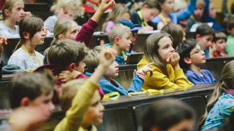 In einem Hörsaal sitzen viele Kinder in den Sitzreihen und hören aufmerksam zu. Einige Kinder heben die Hand, um sich zu melden.