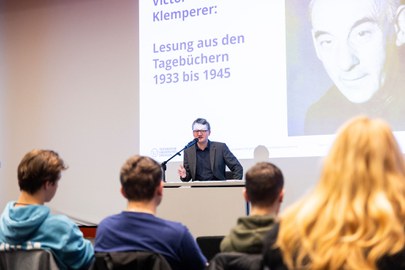 Ein Mann sitzt auf einer Bühne und spricht zu den Menschen, die vor ihm sitzen. 