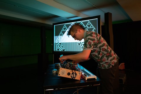 Ein Mann steht vor einer Leinwand und macht Musik an einem DJ-Pult.