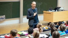 Ein Mann spricht in einem Hörsaal zu Kindern. Die Kinder sitzen auf Hörsaalgestühl mit Schreibplatte und schauen ihn an.