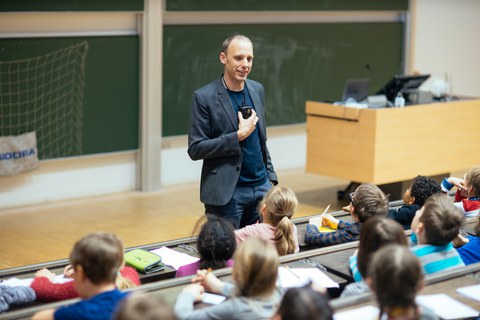Ein Mann spricht in einem Hörsaal zu Kindern. Die Kinder sitzen auf Hörsaalgestühl mit Schreibplatte und schauen ihn an.