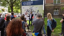 Eine Gruppe von Menschen steht vor einem Gebäude und einer großen Campuskarte der TUD. Im Hintergrund ein Baum.