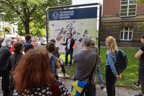 Eine Gruppe von Menschen steht vor einem Gebäude und einer großen Campuskarte der TUD. Im Hintergrund ein Baum.