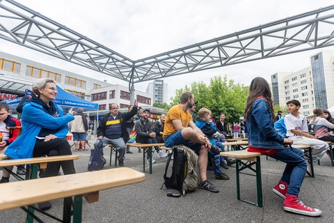 Das Foto zeigt Personen, die auf Festzeltbänken sitzen. Die Bänke stehen in mehreren Reihen hintereinander. Eine der Personen, die dort sitzen, meldet sich. Eine andere Person im Publikum macht mit einer Geste auf die Person, die sich meldet, aufmerksam.  