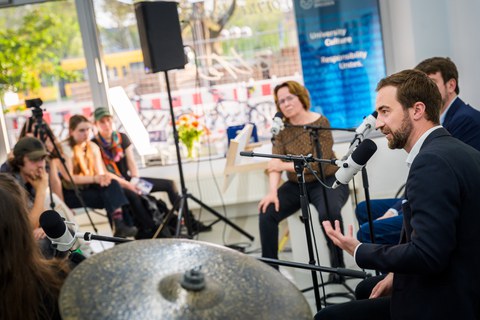 The photo shows 2 men and 1 woman sitting at microphones on the right. One of the men is speaking to people sitting in front of them (on the left of the picture). In the background a large window front.