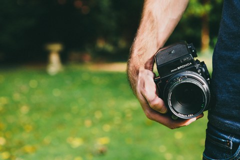 Foto zeigt einen Mann im Grünen, der eine Kamera mit Objektiv in der linken Hand hält.