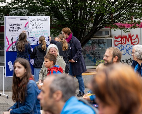 Im Vordergrund des Fotos sitzen Personen verschiedenen Alters. Der Fokus ist auf den Hintergrund gerichtet. Dort stehen 3 Jugendliche und eine erwachsene Person an einer blauen Pinnwand, an der groß die Frage „Was wären wir ohne Freiheit?“ steht.