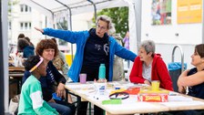 Das Bild zeigt 3 erwachsene Personen und einen Jungen, die an einem Tisch sitzen. Am Tisch steht eine Person mit zur Seite ausgestreckten Armen.