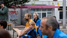 Das Bild zeigt Kinder und Erwachsene, die auf Festzeltbänken sitzen. Im Hintergrund sind mit Graffiti beschmierte Wände zu sehen.