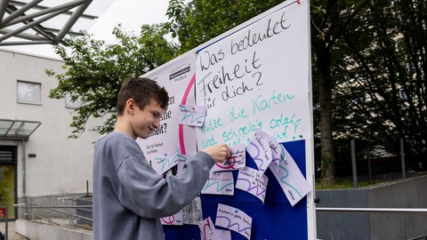 Ein Jugendlicher steht vor einer Pinnwand und steckt eine Karte an der Wand fest. An der Wand steht "Was bedeutet Freiheit für dich?". 