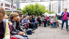Erwachsene, Kinder und Jugendliche sitzen auf Festzeltbänken und hören einem Mann, der in ein Mikro spricht, zu.