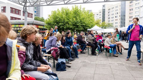 Erwachsene, Kinder und Jugendliche sitzen auf Festzeltbänken und hören einem Mann, der in ein Mikro spricht, zu. 