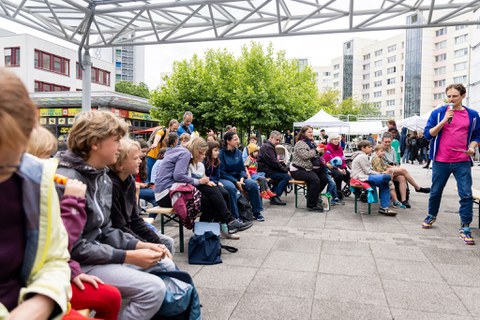 Erwachsene, Kinder und Jugendliche sitzen auf Festzeltbänken und hören einem Mann, der in ein Mikro spricht, zu. 