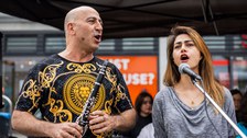 Ein Mann, der eine Klarinette hält, und eine Frau stehen an einem Mikro und singen.