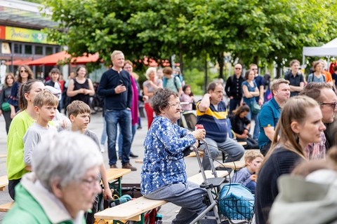 Menschen unterschiedlichen Alters sitzen auf Bänken, die hintereinander stehen. Im Hintergrund stehen Menschen und schauen in dieselbe Richtung wie die sitzenden Menschen. 