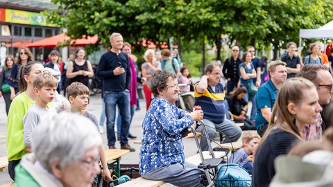 Menschen unterschiedlichen Alters sitzen auf Bänken, die hintereinander stehen. Im Hintergrund stehen Menschen und schauen in dieselbe Richtung wie die sitzenden Menschen. 