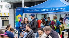 Menschen sitzen und stehen zusammen. Im Hintergrund steht ein Pavillon mit der Aufschrift „TU Dresden | Universitätskultur“.