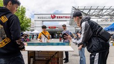 2 erwachsene Männer und 3 Jugendliche spielen Tischtennis mit ungewöhnlichen Schlägern auf einer Platte, die aus verschiedenem Material besteht.