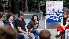 Das Foto zeigt 3 Personen, die auf Festzeltbänken sitzen. Die Person rechts hält ein Mikro und spricht. Im Hintergrund ist eine blaue Pinnwand zu sehen, an der ein Poster mit der Frage "Was wären wir ohne Freiheit?" hängt.