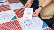 Das Foto zeigt einen Festzelttisch, auf dem Karten liegen. Bei manchen Karten ist die Vorderseite mit einer Zeichnung zu sehen. Von anderen Karten sieht man nur die rote Rückseite mit weißer Schrift. Eine Person hält das Grundgesetz der BRD in die Kamera.