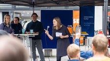 Eine Person mit Mikro schaut und zeigt zu Personen, die auf dem Bild nicht zu sehen sind. Im Hintergrund stehen links 3 Personen an einem Stehtisch neben einer blauen Pinnwand.
