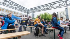Das Foto zeigt Personen, die auf Festzeltbänken sitzen. Die Bänke stehen in mehreren Reihen hintereinander. Eine der Personen, die dort sitzen, meldet sich. Eine andere Person im Publikum macht mit einer Geste auf die Person, die sich meldet, aufmerksam.