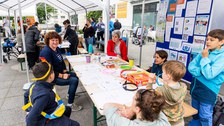 Erwachsene und Kinder sitzen und stehen an einem Tisch unter einem Pavillon. Rechts im Bild steht eine blaue Pinnwand, an der verschiedene Zettel hängen.