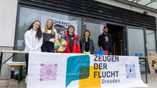 6 Personen stehen hinter einem Tisch, an dem ein Banner mit dem Aufdruck "Zeugen der Flucht2 trägt