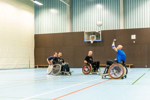 Foto mehrerer Männer in einer Turnhalle. Die Männer sitzen in Rollstühlen und spielen Rugby. 