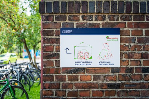 Foto einer Backstein mit einem Schild. Das Schild ist ein Wegweiser des Campusbüro „Uni mit Kind“ zum Aufenthaltsraum und zum Wickelraum