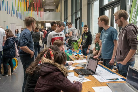 Foto im Foyer des Neubaus der Chemischen Institute zum dies academicus. Es sind viele Personen an Informationsständen zu sehen. Von der Decke hängen Stäbe in den Spektralfarben.