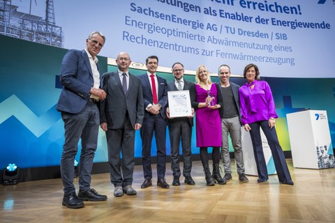 Fotoaufnahme des Gewinnerteams während der Preisverleihung auf der Bühne mit den Moderierenden von BMWK (links) und ARD/BR (rechts)