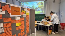 Foto eines Seminarraums. Der linken Teil des Fotos wird von einer Tafel eingenommen, die mit orangenen, beschriebenen Zetteln behängt ist und schräg nach hinten in den Raum hineinragt. Rechts im Hintergrund befindet sich eine Gruppe von Personen, die um einen Tisch herum steht, beziehungsweise sitzt.