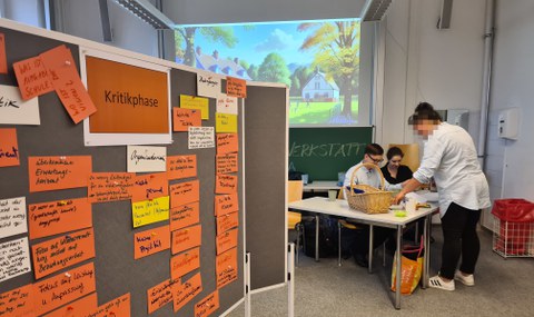 Foto eines Seminarraums. Der linken Teil des Fotos wird von einer Tafel eingenommen, die mit orangenen, beschriebenen Zetteln behängt ist und schräg nach hinten in den Raum hineinragt. Rechts im Hintergrund befindet sich eine Gruppe von Personen, die um einen Tisch herum steht, beziehungsweise sitzt.