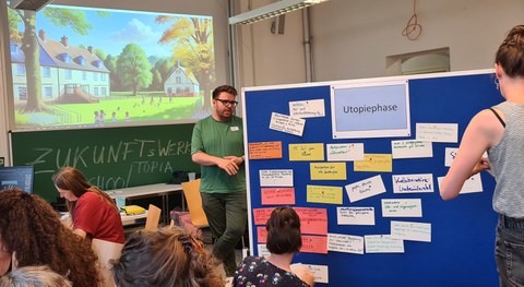 Foto eines Seminarraums. Der rechte Teil des Fotos wird von einer Tafel eingenommen, die mit orangenen, beschriebenen Zetteln behängt ist. Darum stehen Personen, die Zettel aufhängen. In der linke Hälfte des Fotos sitzen Personen um einen Tisch herum. Im Hintergrund ist eine Tafel zu sehen. Darüber wird ein Bild einer Landschaft mit Häusern projiziert.
