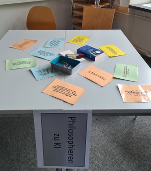 Das Foto zeigt einen quadratischen Tisch von schräg oben. Auf dem Tisch liegen bunte Zettel mit Texten, Stifte und Reißzwecken in einer Dose. An die vordere Kante ist ein Blatt Papier geheftet, auf dem „Philosophieren zu KI“ zu lesen ist.