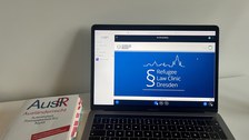Foto eines aufgeschlagenen Laptops auf einem Tisch, daneben liegt ein Buch. Auf dem Laptop ist eine blaues Bild mit dem Text "Refugee Law Clinic Dresden".
