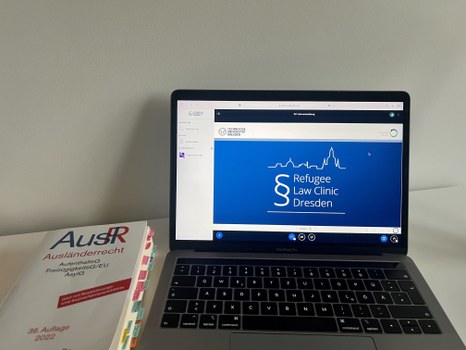 Foto eines aufgeschlagenen Laptops auf einem Tisch, daneben liegt ein Buch. Auf dem Laptop ist eine blaues Bild mit dem Text "Refugee Law Clinic Dresden".