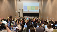 Das Foto zeigt einen Hörsaal mit Blick auf die Tafel. Im Vordergrund sitzen viele Personen mit dem Rücken zum Betrachter. Sie halten verschiedenfarbige Zettel in die Luft. Vor jeder Person steht ein Laptop. Im Hintergrund wird ein Bild an die Wand projiziert, welches 4 verschiedenfarbige Quadrate zeigt.