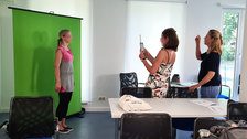 Foto zweier Menschen. Die Person links steht vor einem Greenscreen, die Person rechts fotografiert die andere Person