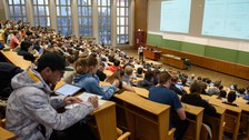 Foto: Voller Hörsaal mit Studierenden, vorne steht ein Mann und zeigt auf die die projizierten Folien