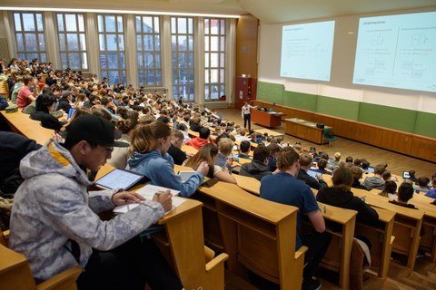Foto: Voller Hörsaal mit Studierenden, vorne steht ein Mann und zeigt auf die die projizierten Folien