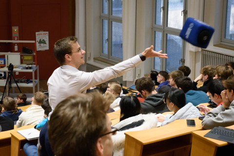 Foto: Hörsaal mit Studierenden, ein Mann steht und wirft ein Wurfmikrofon (Würfel)