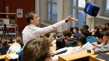 Foto: Hörsaal mit Studierenden, ein Mann steht und wirft ein Wurfmikrofon (Würfel) 