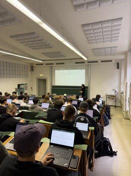 Foto: Kleiner Hörsaal, man sieht von hinten viele Studierende mit ihren Laptop, vorne an der Tafel steht ein Mann und 
