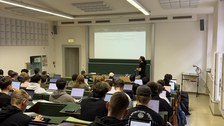 Foto: Kleiner Hörsaal, man sieht von hinten viele Studierende mit ihren Laptop, vorne an der Tafel steht ein Mann und 