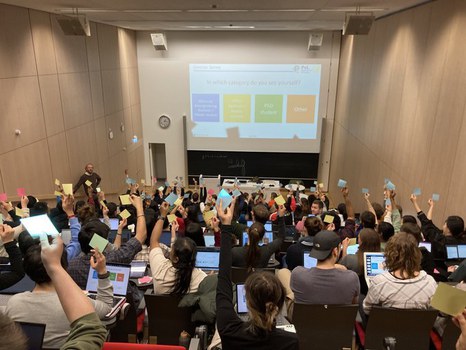 Das Foto zeigt einen Hörsaal mit Blick auf die Tafel. Im Vordergrund sitzen viele Personen mit dem Rücken zum Betrachter. Sie halten Zettel in die Luft. Im Hintergrund wird ein Bild an die Wand projiziert, welches 4 verschiedenfarbige Quadrate zeigt.