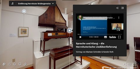 Die Abbildung zeigt eine Screenshot. Im Hintergrund ist ein weißer Raum mit ausgestellten Musikinstrumenten zu erkennen. Auf der rechten Hälfte der Abbildung befindet sich ein dunkelgraues Quadrat, darin ist ein YouTube-Video eingebettet. Darunter steht "Sprache und Klang - die Herrnhuterische Liedüberlieferung".